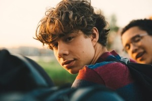 Close-up of a teenage boy sitting outside with friends, looking at the camera.