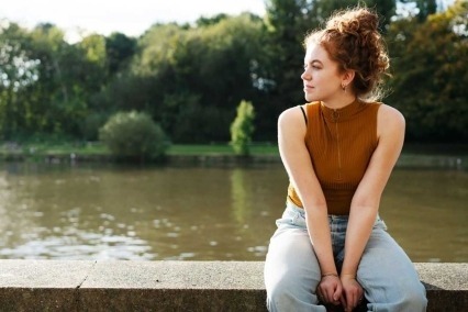 Mujer joven afuera en el sol sentada en una pared al lado del río.
